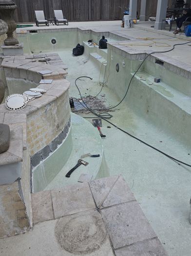 Drained pool with tile removed along a curved wall and hand tools on the floor during renovation